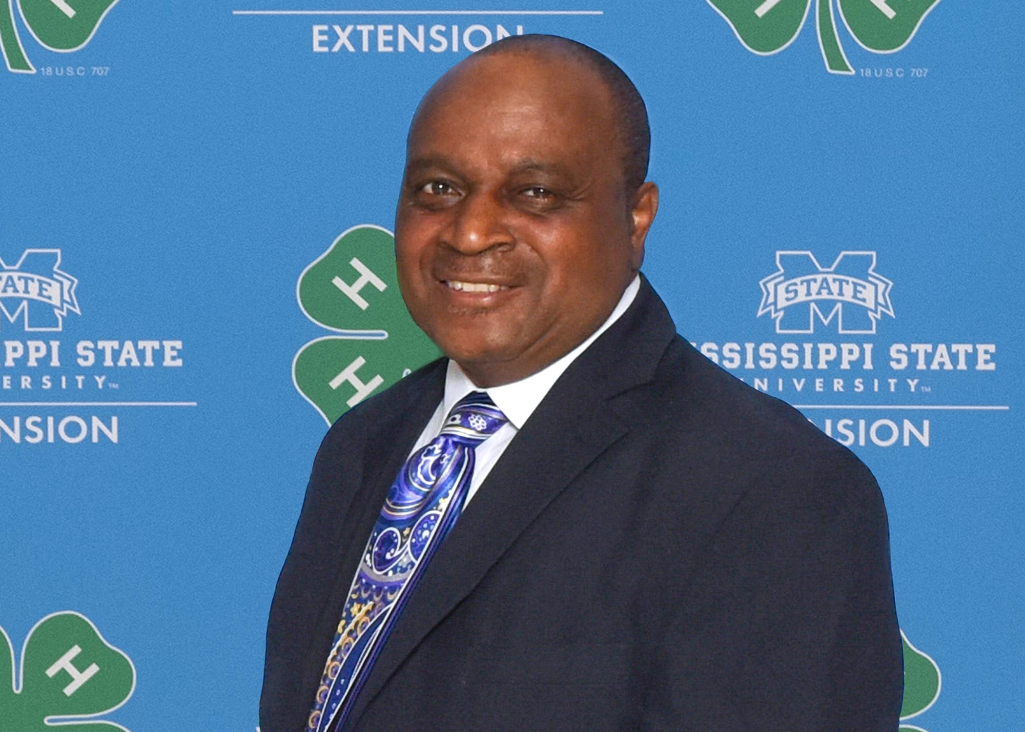 An African American man in a suit stands in front of a light blue background with the MSU Extension Service logo repeated