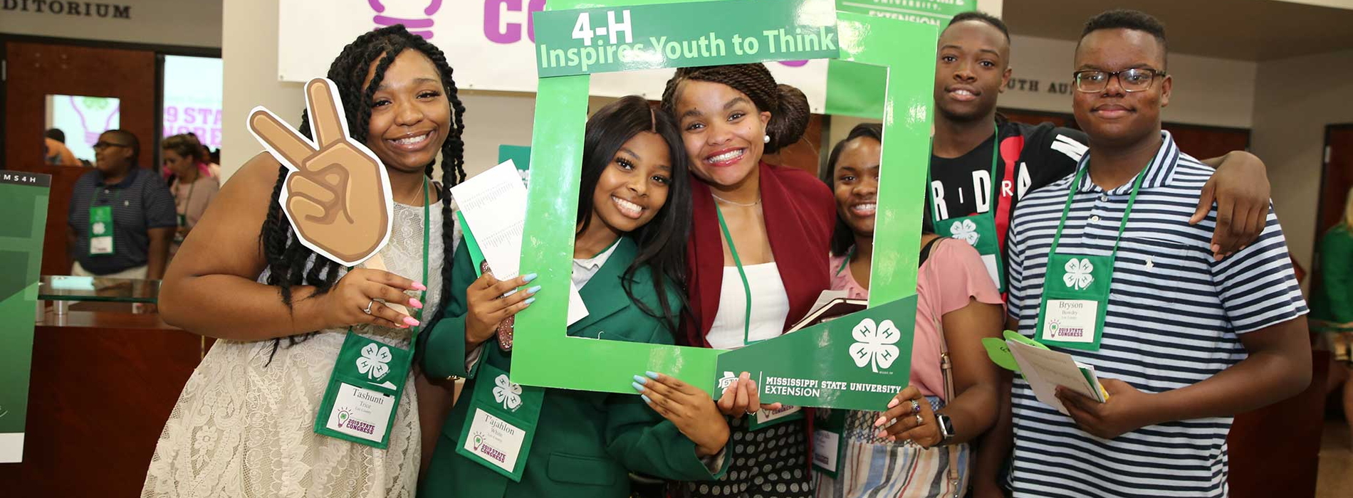 Six 4-H youth take a picture behind a green 4-H photo frame