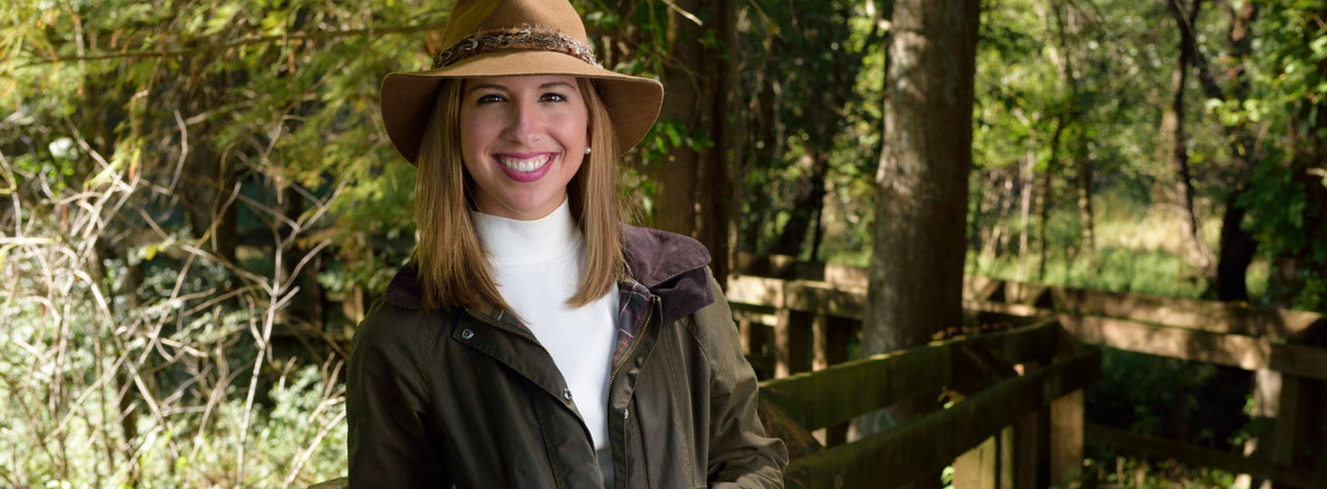 Ashley Ward, Former 4-H member A young woman with brown hair wearing a tan hat with a green jacket and khaki pants leans against a wooden railing.
