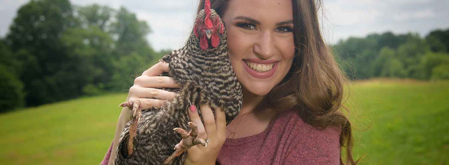 Chicken Care A young woman with brown hair and a pink shirt stands smiling while holding a black and white speckled chicken next to her face.