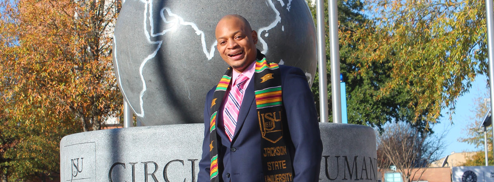 A man wearing a Jackson State University scarf and a navy suit jacket smiles in front of a sculpture on the JSU campus.