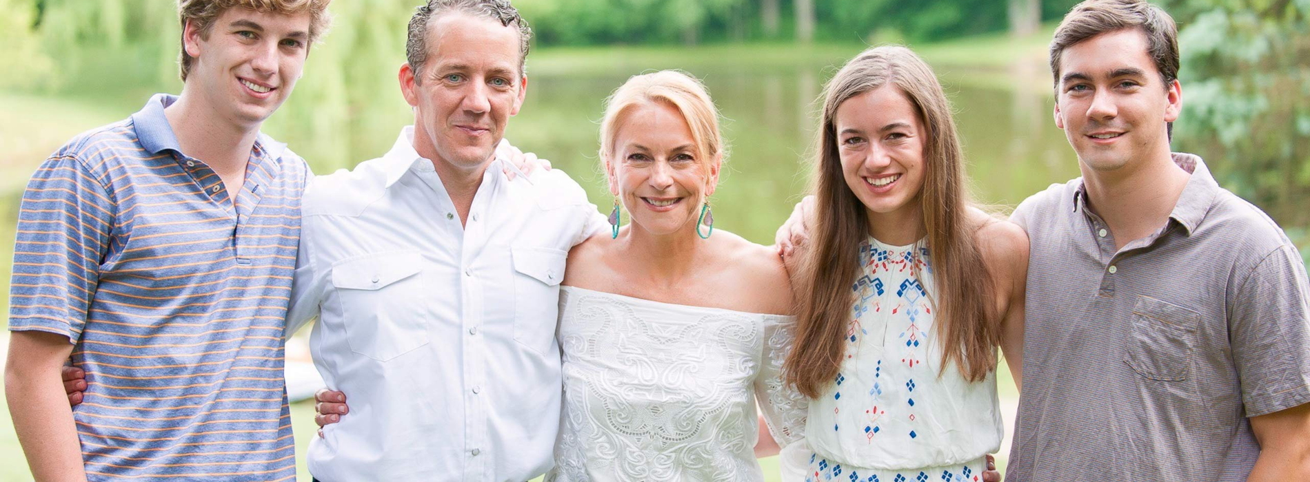 Family photo of 2 parents and 3 young adults standing with arms around each other in front of a small body of water.