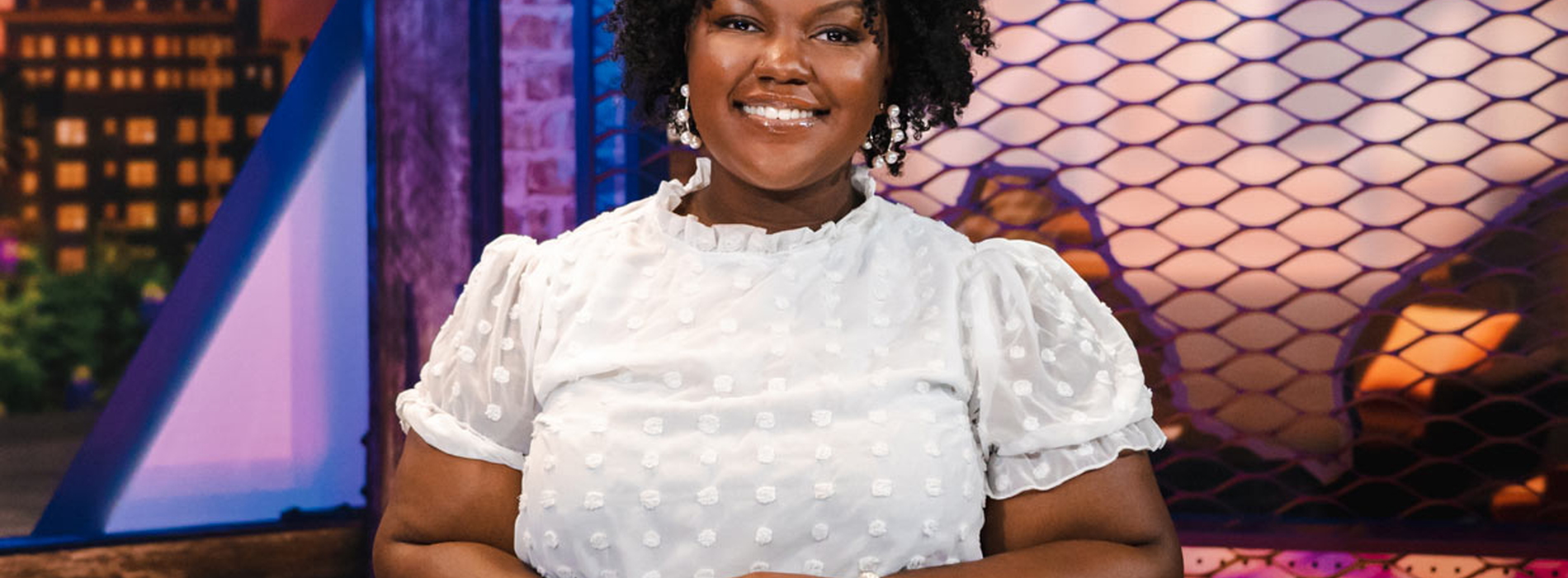 A smiling woman wearing a white shirt sits behind a desk with her hands resting on the desk in front of her.