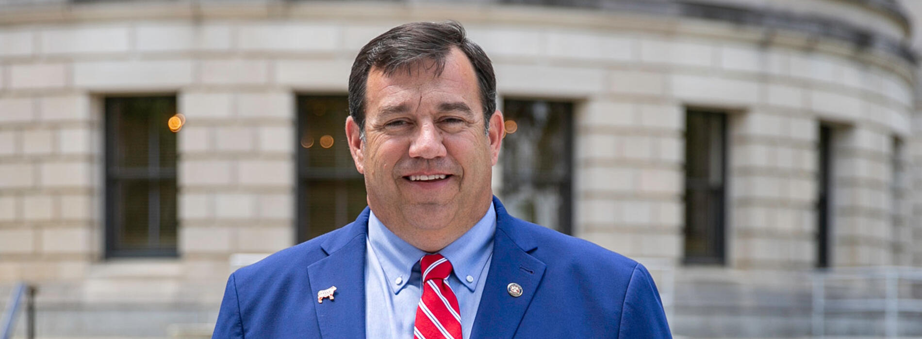A man, smiling, standing outside and wearing a blue suit with a white-striped red tie.