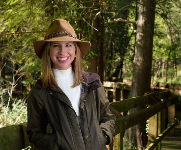 A young woman with brown hair wearing a tan hat with a green jacket and khaki pants leans against a wooden railing.