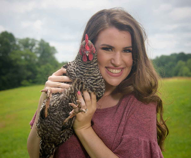 A young woman with brown hair and a pink shirt stands smiling while holding a black and white speckled chicken next to her face. 