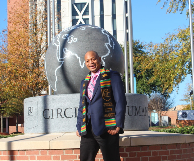 A man wearing a Jackson State University scarf and a navy suit jacket smiles in front of a sculpture on the JSU campus.