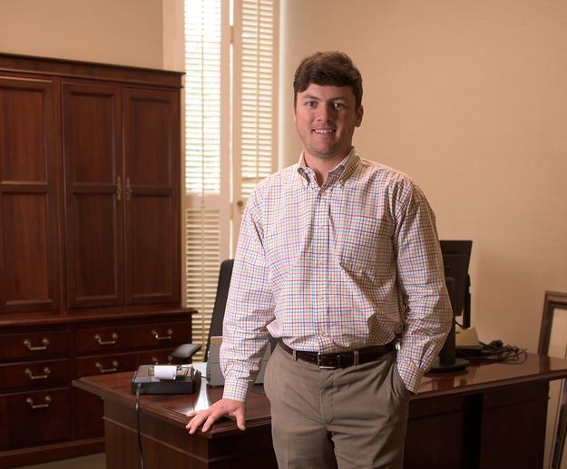 A man wearing a colorful plaid shirt stands in front of a desk.