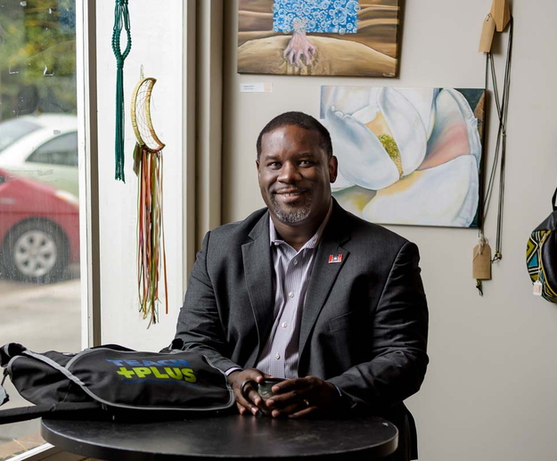 A man wearing a suit sits at a round table with a small mug and a bag that says “Teach Plus.”