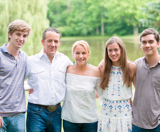 Family photo of 2 parents and 3 young adults standing with arms around each other in front of a small body of water.