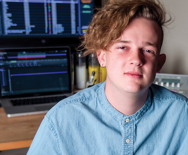 A young man sitting at a desk with computer monitors behind him.