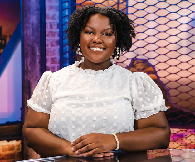 A smiling woman wearing a white shirt sits behind a desk with her hands resting on the desk in front of her.