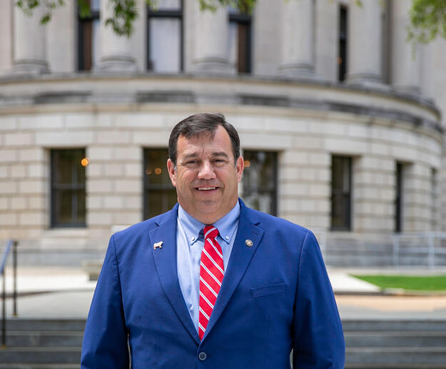 A man, smiling, standing outside and wearing a blue suit with a white-striped red tie.